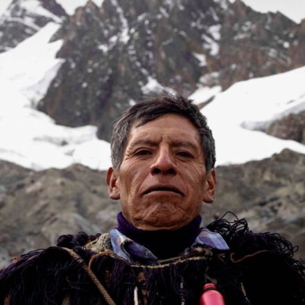 Older Andean man in wooly robe standing before disappearing glacier in the high Andes